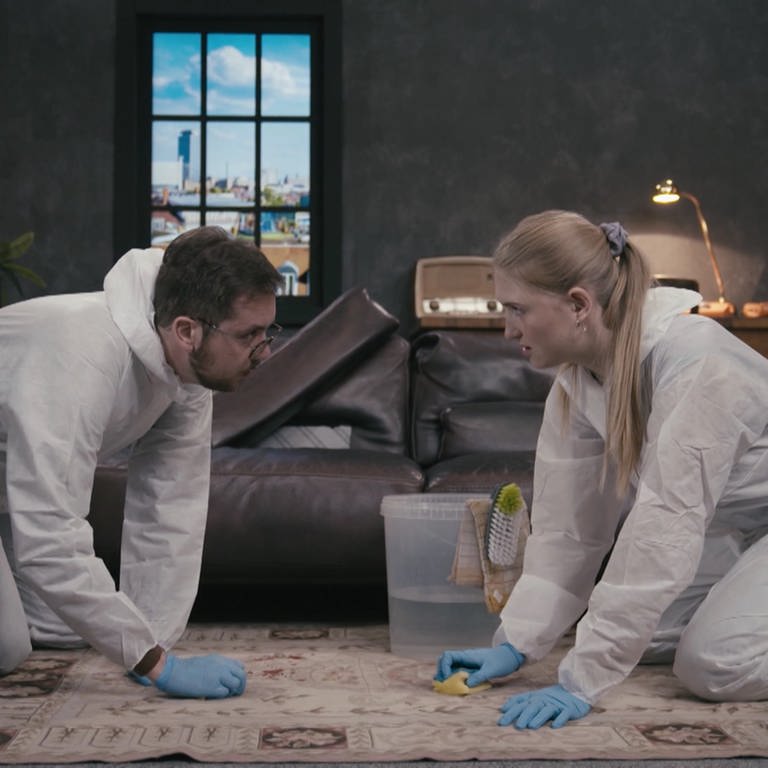Zwei Personen in Schutzanzügen knien auf einem Teppich und reinigen ihn mit Schwämmen.