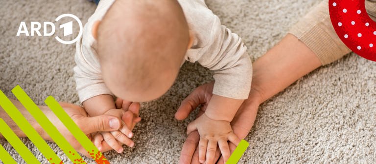 Ein Baby auf dem Boden greift nach den Händen seiner Eltern.