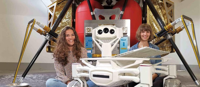 neuneinhalb-Reporterin Mona und Wissenschaftlerin Nadine sitzen vor einer Raumsonde, neben ihnen steht ein Rover, der eines Tages auf dem Mond umherfahren soll.