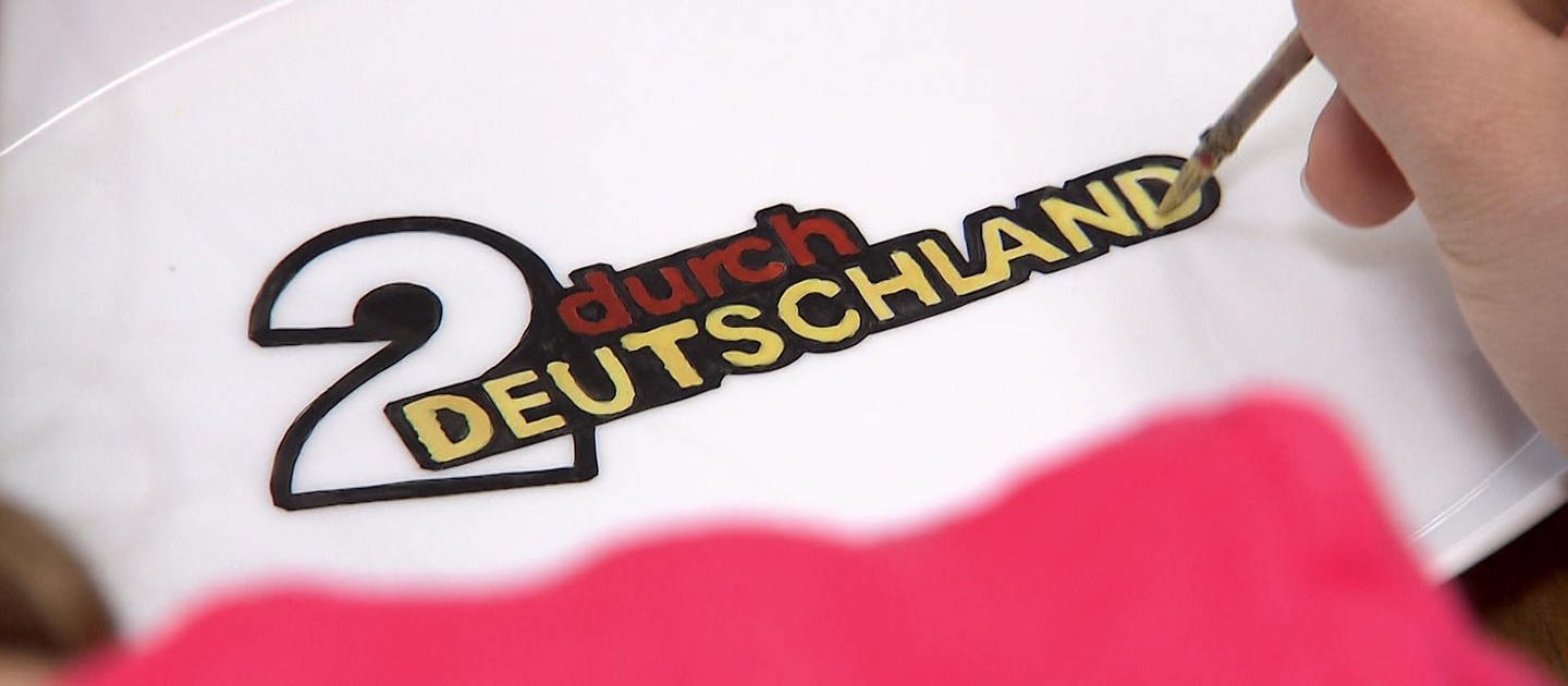 Eine Hand malt mit einem Pinsel das Logo von "2 durch Deutschland" auf Porzellan.