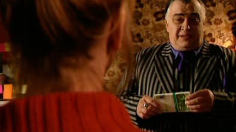 Ein korpulenter Mann im grau-gestreiften Anzug hält ein Bündel mit grünen Geldscheinen in der Hand. Vor ihm, nur von hinten zu sehen, eine Frau in roter Strickweste.