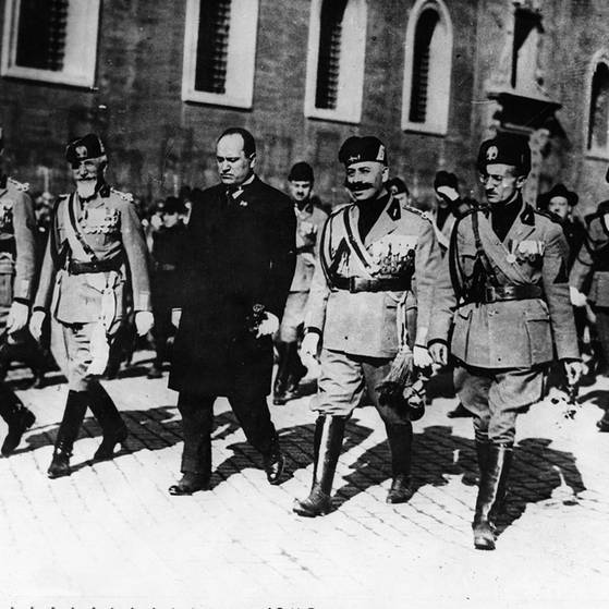 Mussolini beim Marsch auf Rom, er trägt einen schwarzen Mantel und läuft in der ersten Reihe, neben und hinter ihm marschieren viele Männer in Uniform.
