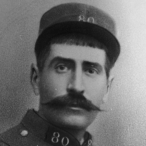 Ein schwarz-weiß Foto von einem Soldaten mit Schnurrbart und Uniform, der in die Kamera schaut.