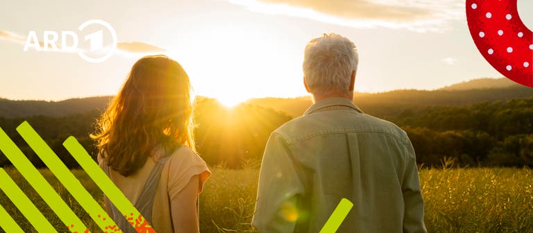 Ein Vater steht mit seiner Tochter in einem Feld und sieht einem Sonnenuntergang zu.