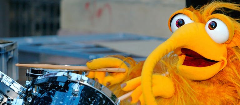 Eine gelbe Plüschpuppe spielt auf einem Schlagzeug.