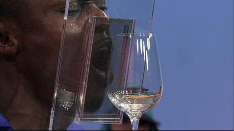 Ein Mann steht vor einer Glasscheibe und singt, dahinter steht ein Weinglas.