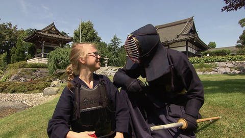 Ein Mann und eine Frau sind in einem japanischen Garten und tragen Samurai-Rüstungen. 