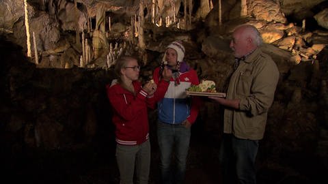 Ein Mann und eine Frau stehen in einer Tropfsteinhöhle und essen Käse. Ein älterer Mann hält ihnen eine Käseplatte hin. 