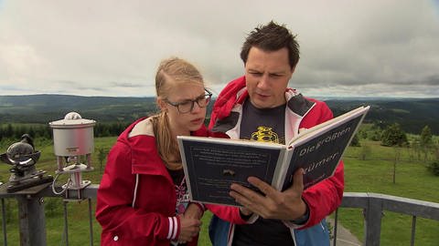 Ein Mann und eine Frau stehen auf einem Berg und lesen im Buch „Die größten Irrtümer im Sauerland“.