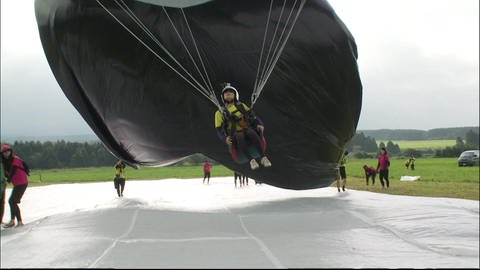 Eine Person hängt an Seilen befestigt an einem Heißluftballon, dieser hebt ab.