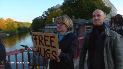 Die Moderatorin und ein Mann spazieren an einem Fluss. Sie hält ein Schild mit der Aufschrift: Free Hugs.