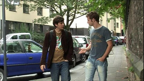 Zwei junge Männer laufen auf der Straße nebeneinander her und sprechen miteinander.