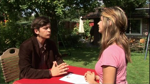 Ein junger Mann und eine junge Frau sitzen im Garten an einem Tisch und simulieren ein Bewerbungsgespräch.