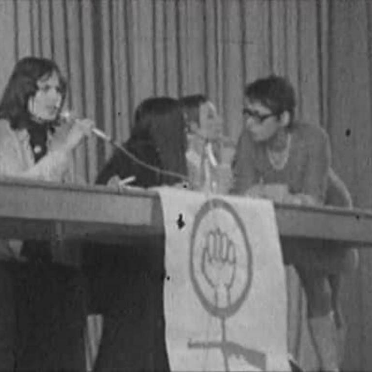Schwarz-weiß Bild von einem langen Tisch auf einer Tribüne. An dem Tisch sitzen fünf Frauen, eine spricht in ein Mikrofon. Es hängt eine Fahne mit geballter Faust vor dem Tisch.