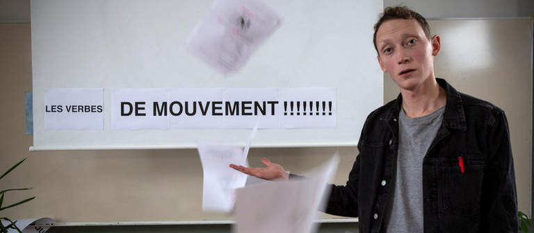 Ein junger Mann steht vor einer Tafel, darauf steht "Les verbes de mouvement!!!!!!". Um ihn herum fliegen Blätter.