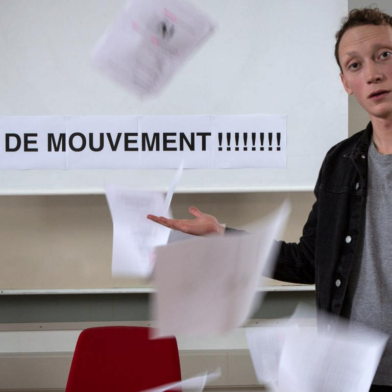 Ein junger Mann steht vor einer Tafel, darauf steht "Les verbes de mouvement!!!!!!". Um ihn herum fliegen Blätter. (Foto: WDR)