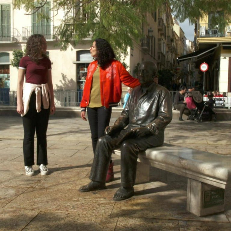 Zwei Frauen stehen auf einem Platz. Eine der beiden legt ihre Hand auf eine Bronzestatue eines Mannes, der auf einer Bank sitzt. (Foto: WDR)