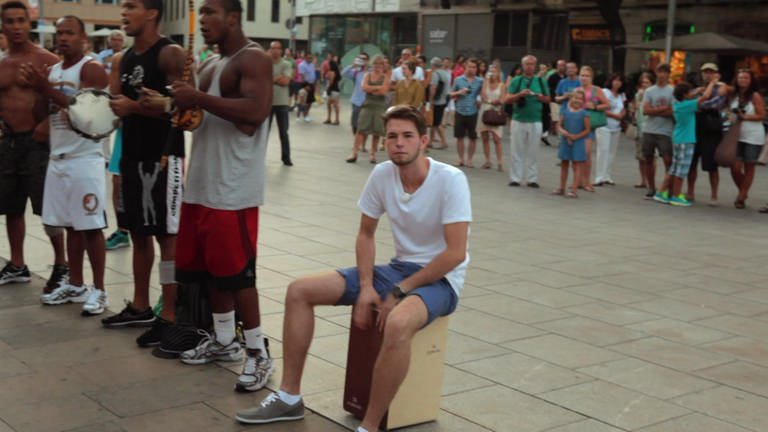 Innenstadt: Ein junger Mann sitzt auf einem Cajón. Um ihn herum sind viele Menschen.
