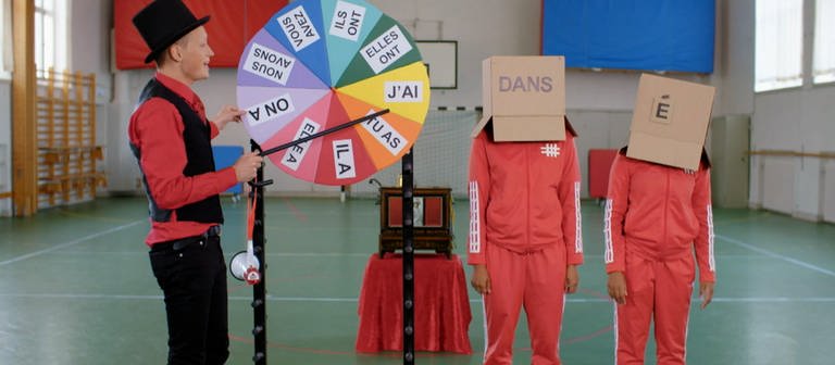 Ein junger Mann steht an einem bunten Glücksrad, hinter ihm zwei Personen mit Kartons auf dem Kopf. Auf einem Karton steht "Dans", auf dem anderen "É".