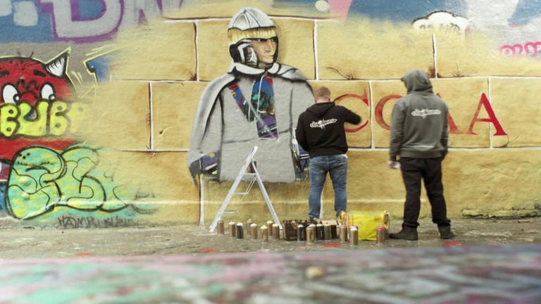 Zwei Männer sprühen Graffiti auf eine gelbe Wand.