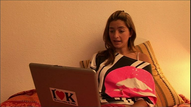 Eine junge Frau liegt im Bett und schaut auf ihren Laptop, sie spricht.