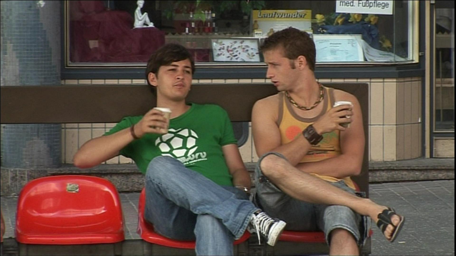 Zwei junge Männer sitzen an einer Bahnhaltestelle, trinken Kaffe und reden miteinander.