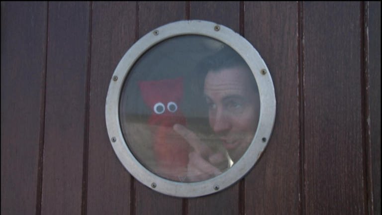 Ein Mann und eine rote Strumpfhandpuppe sind auf einem Schiff und schauen durch ein Bullauge.