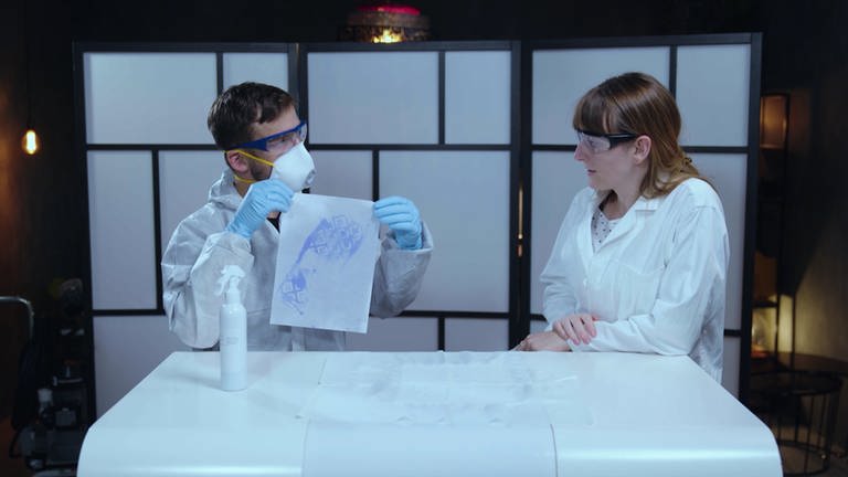 Zwei Menschen in Laborkitteln mit einem Blatt Papier, auf dem ein Schuhabdruck durch Besprühen mit einer chemischen Lösung sichtbar gemacht wurde.