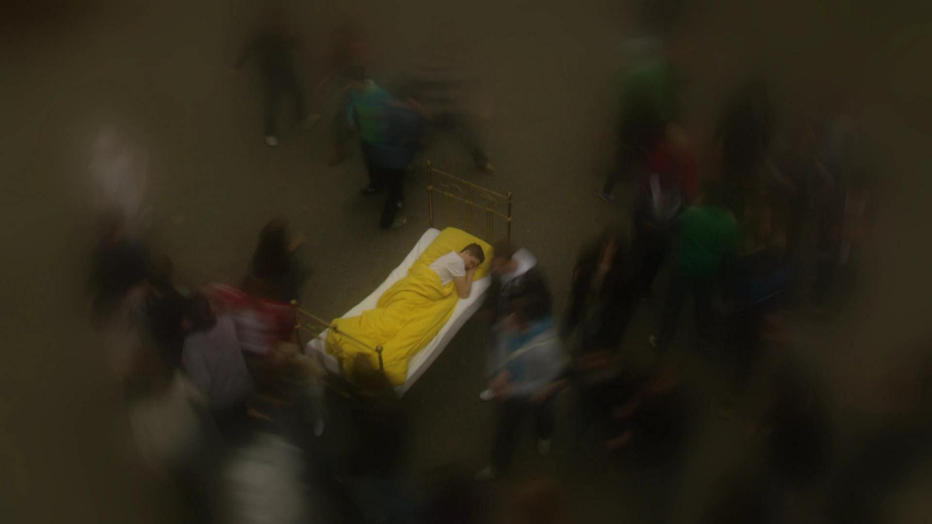 Ein Mann schläft in einem Bett auf einem Platz. Um ihn herum laufen Menschen, die verschwommen abgebildet sind. (Foto: WDR)