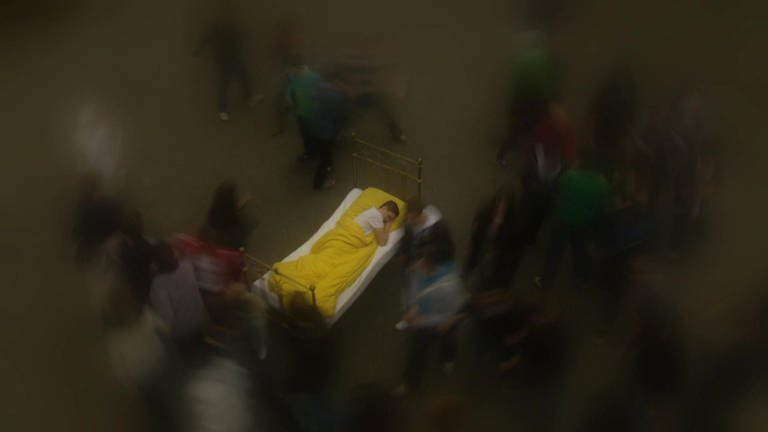 Ein Mann schläft in einem Bett auf einem Platz. Um ihn herum laufen Menschen, die verschwommen abgebildet sind.