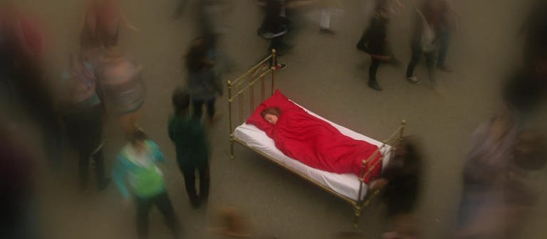 Eine Frau schläft in einem Bett auf einem Platz. Um sie herum laufen Menschen, die verschwommen abgebildet sind. (Foto: WDR)