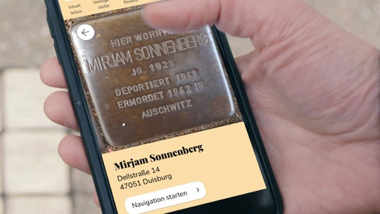 Ein Handy zeigt die Stolpersteine-App des WDR mit dem Bild eines Stopersteines aus Metall. Seine Aufschrift: Hier wohnte Mirjam Sonnenberg, Jg. 1925. Deportiert 1943, ermordet 1942 in Auschwitz. (Foto: WDR)
