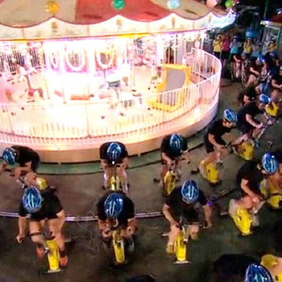 Radfahrer auf Ergometern vor einem beleuchteten Karussell. (Foto: SWR – Screenshot aus der Sendung)
