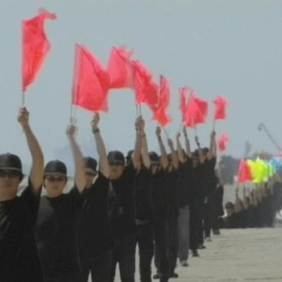 Eine Reihe dunkel gekleideter Menschen hebt rote Flaggen hoch. (Foto: SWR - Screenshot aus der Sendung)