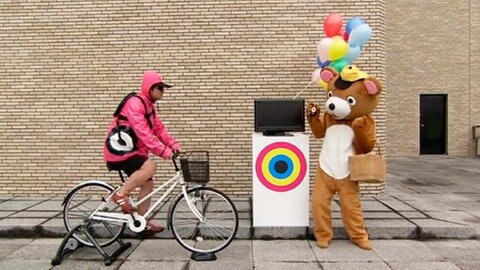 Radfahrer vor einem Fernseher, daneben ein Mensch im Bärenkostüm.