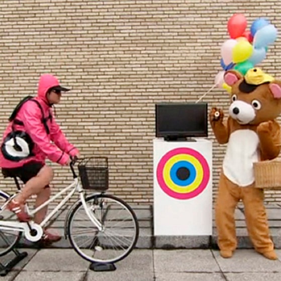 Radfahrer vor einem Fernseher, daneben ein Mensch im Bärenkostüm.