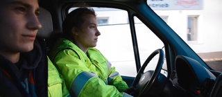 Eine junge Frau und ein junger Mann sitzen im Auto. Beide tragen neongelbe Arbeitsjacken.