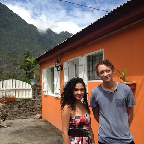 Eine junge Frau und ein Mann vor einem orangefarbenen Haus; im Hintergrund ein Berg. (Foto: WDR)