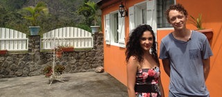Eine junge Frau und ein Mann vor einem orangefarbenen Haus; im Hintergrund ein Berg.