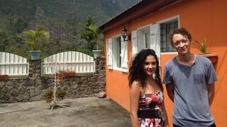 Eine junge Frau und ein Mann vor einem orangefarbenen Haus; im Hintergrund ein Berg.