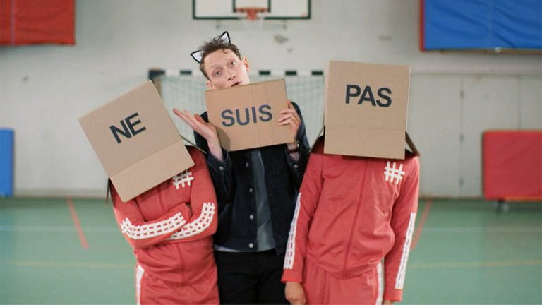 Ein junger Mann steht zwischen zwei Personen mit Karton auf dem Kopf, es bildet sich der Ausdruck "ne suis pas"