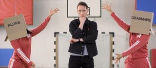 Ein junger Mann steht zwischen zwei Personen, die Kartons auf dem Kopf tragen. Darauf steht "Alors Grammaire".