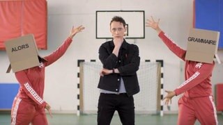 Ein junger Mann steht zwischen zwei Personen, die Kartons auf dem Kopf tragen. Darauf steht "Alors Grammaire".