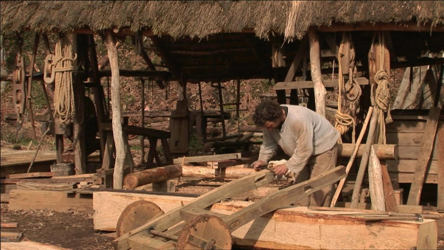 Ein Mann bearbeitet einen kleinen Wagen aus Holz mit einem Ziehmesser. loading=
