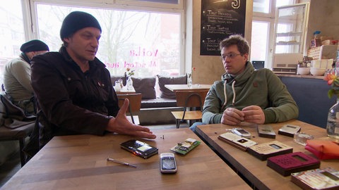 Zwei Männer an einem Tisch, vor ihnen selbst gebaute Handys.