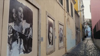 Eine schmale Gasse mit großen Bildern von Personen an der linken Hauswand.