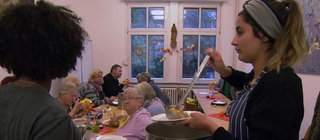 Im Hintergrund sitzen mehrere ältere Menschen an einem Tisch. Im Vordergrund verteilen junge Menschen Essen.