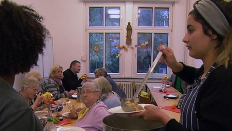 Im Hintergrund sitzen mehrere ältere Menschen an einem Tisch. Im Vordergrund verteilen junge Menschen Essen.