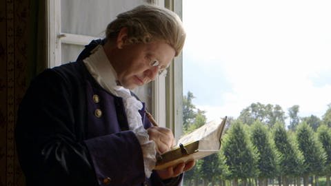 Mann mit Perücke schreibt in ein Buch. (Foto: SWR/WDR – Screenshot aus der Sendung)