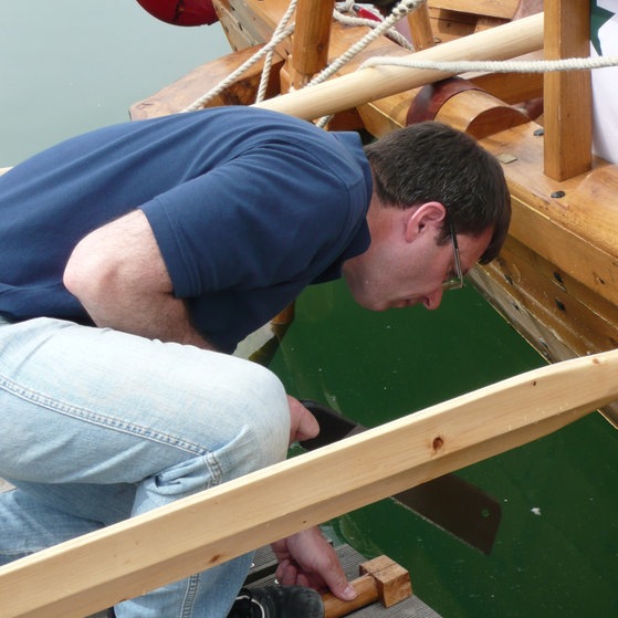 Ein Mann beugt sich vor dem nachgebauten Römerschiff ans Wasser.